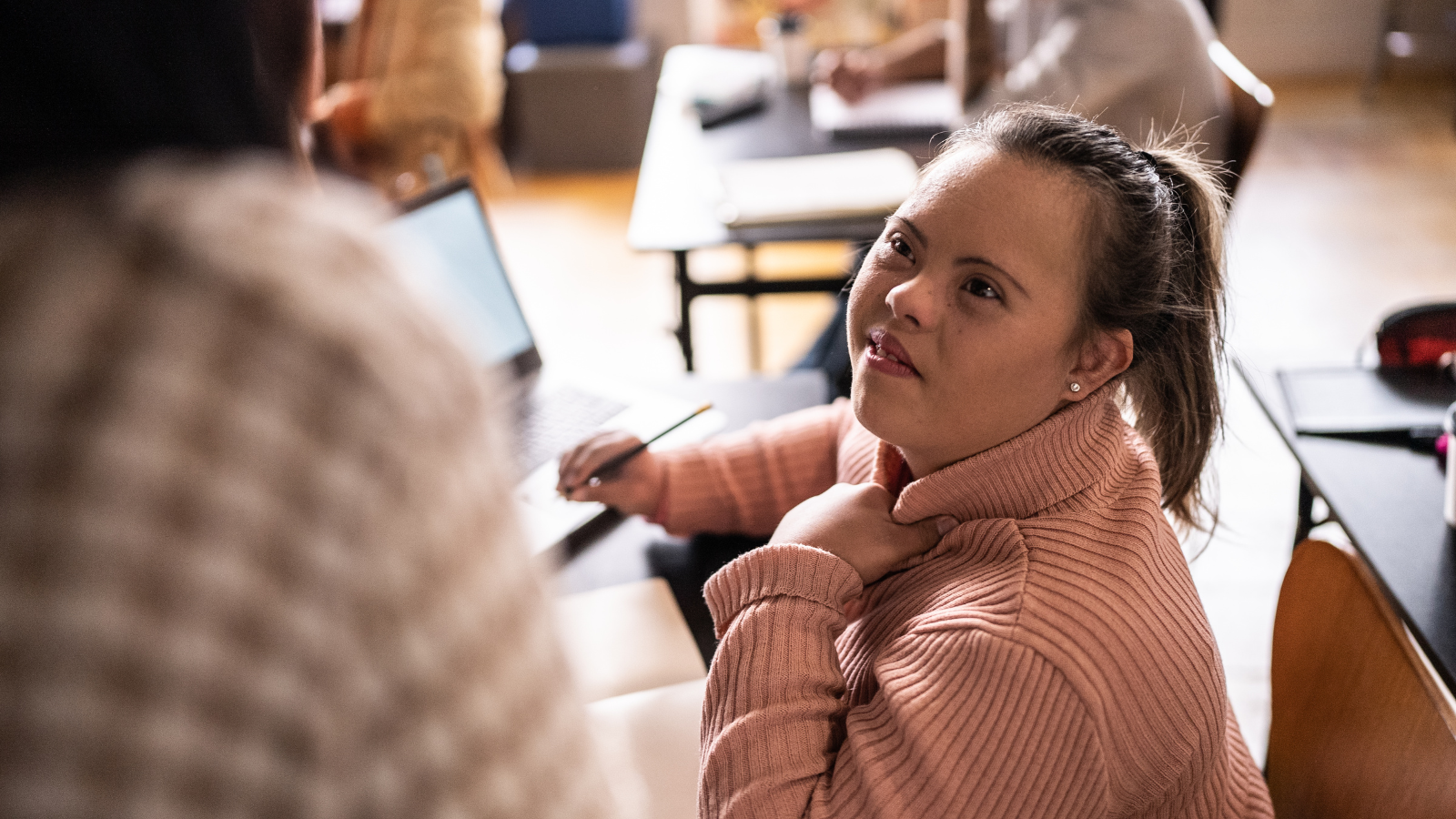 A young woman with Down syndrome sits at a desk in a classroom, looking up attentively at someone speaking to her. She is wearing a pink turtleneck sweater and holding a pencil, with an open notebook and laptop in front of her. The setting appears to be a warm and supportive learning environment.